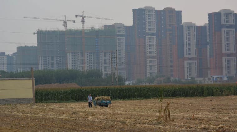 A farmer in the foreground in a hayfield, buildings being constructed in the background.
